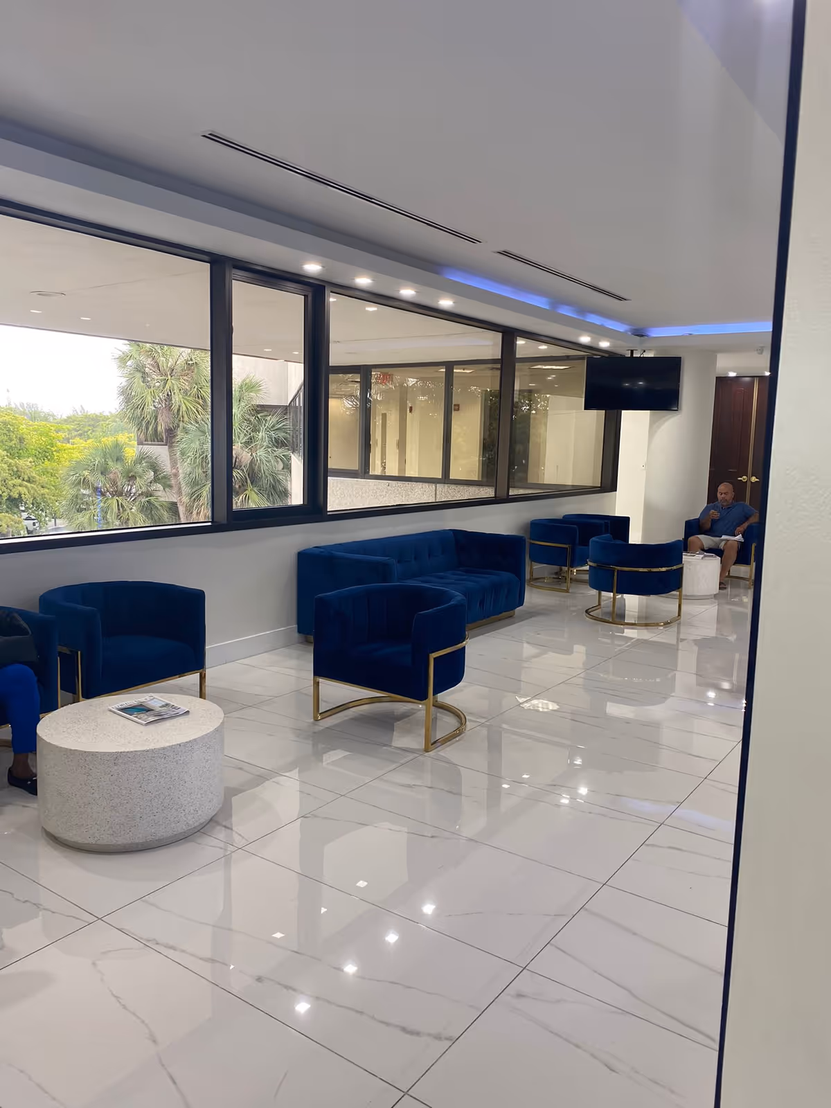 Modern lounge with blue velvet chairs, glass windows, and a seated man.