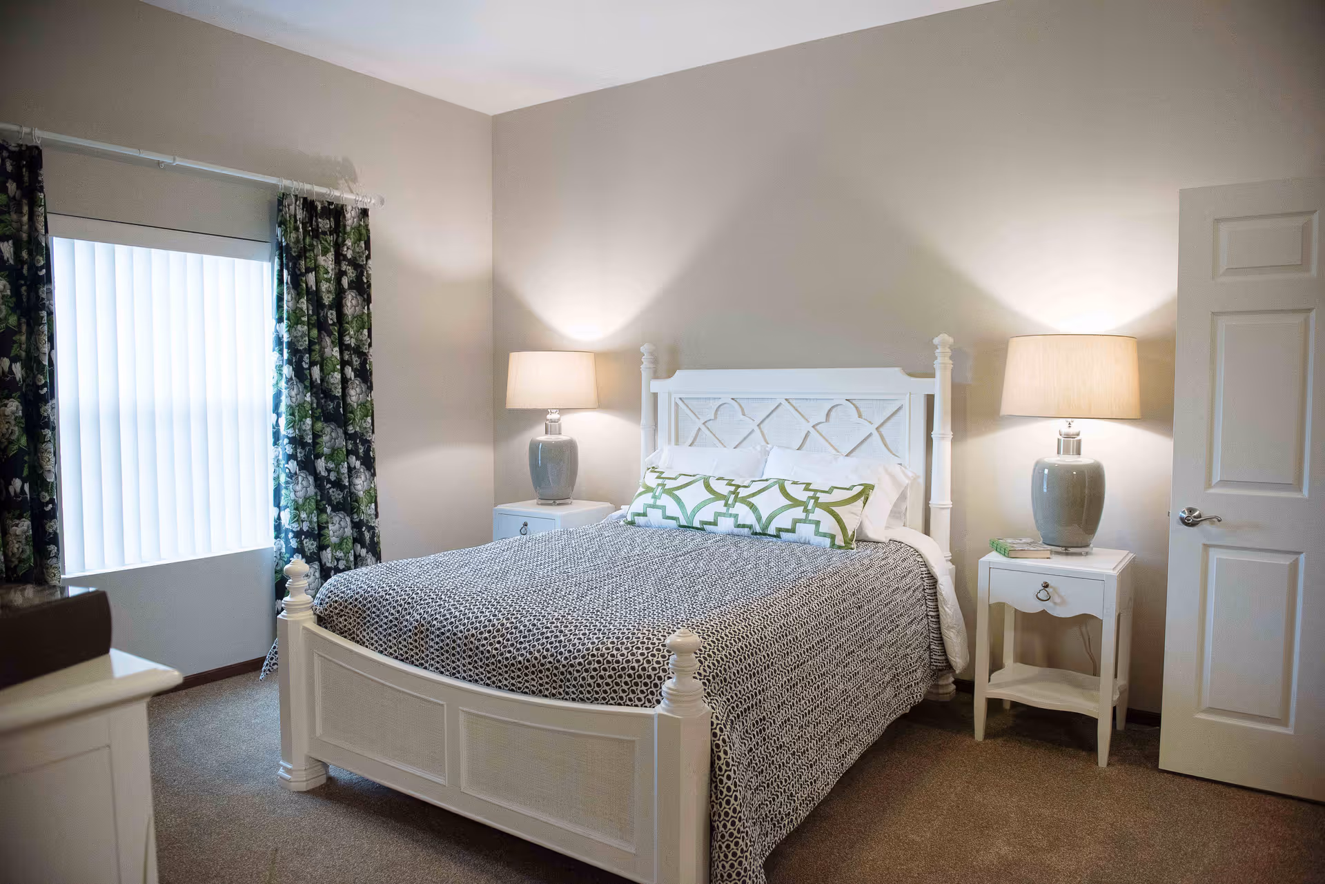 A bedroom with a white wooden bed frame, a patterned gray and white bedspread, and two green and white decorative pillows. There are two matching white nightstands on either side of the bed, each with a gray lamp with a beige lampshade. A window with floral curtains is on the left wall, and a white door is partially open on the right side of the room.
