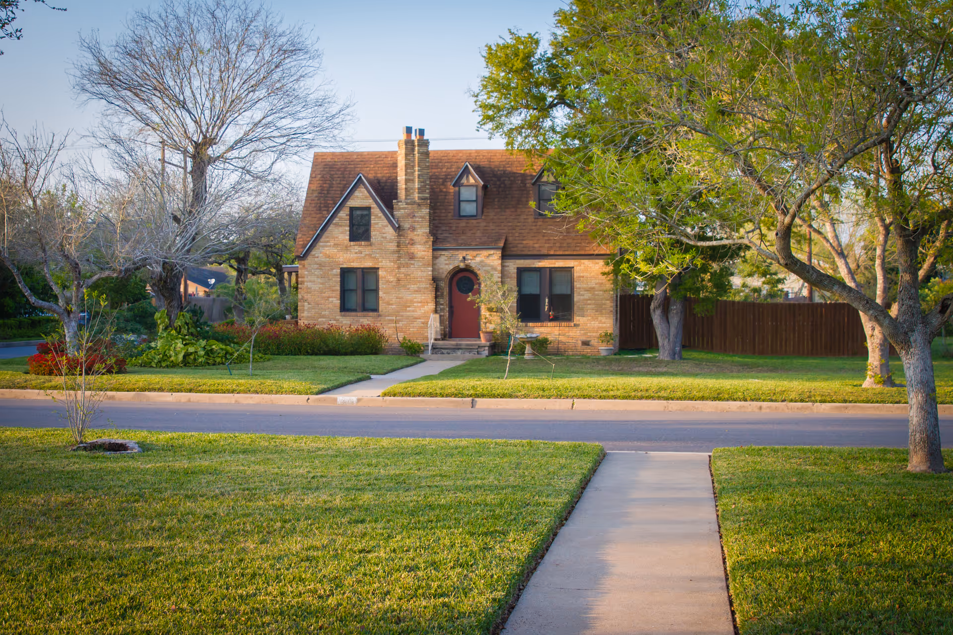 A brick house with a manicured front yard, trees, and a sidewalk.