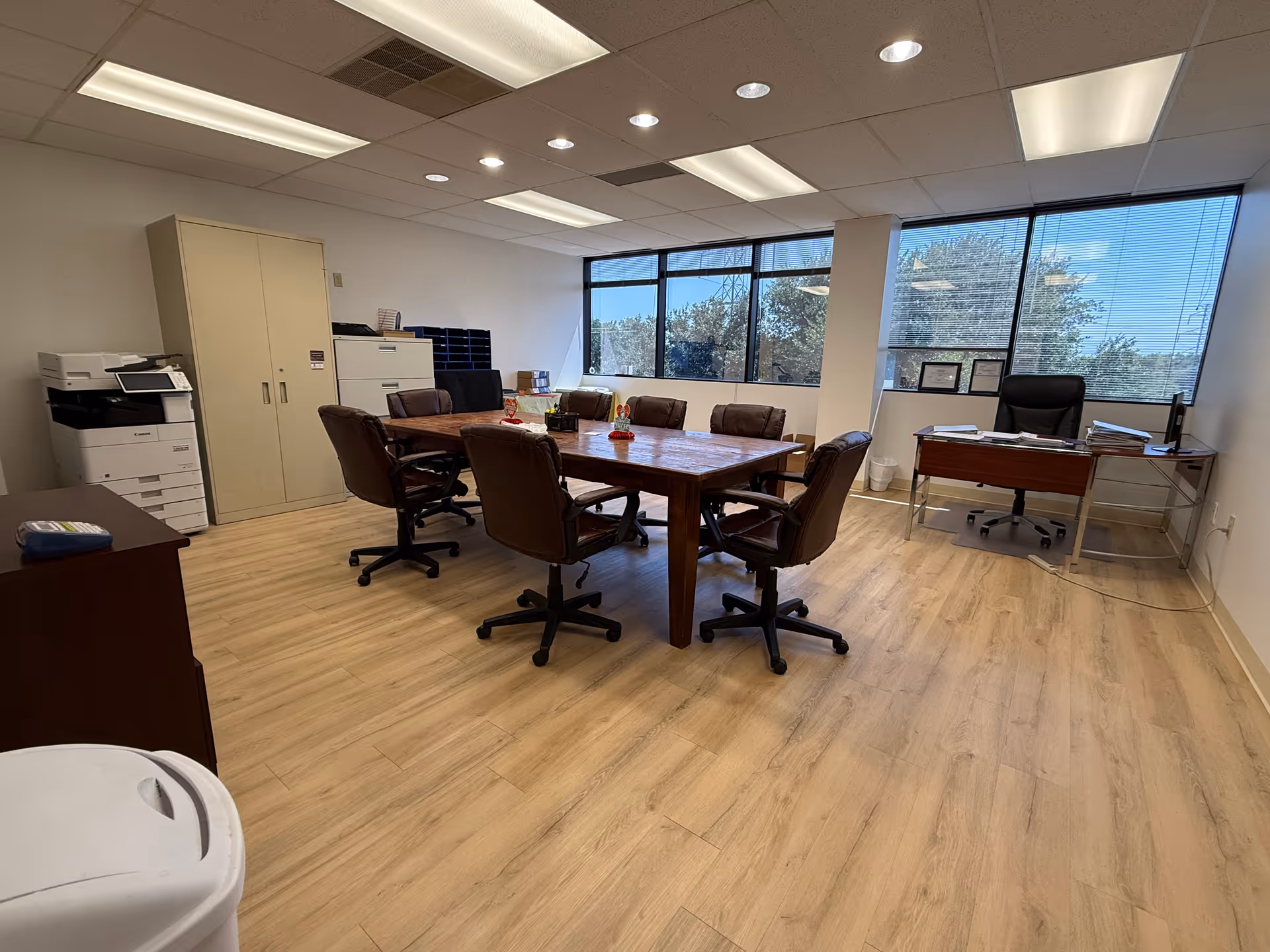 A conference room with a wooden table, brown chairs, large windows, and office equipment.