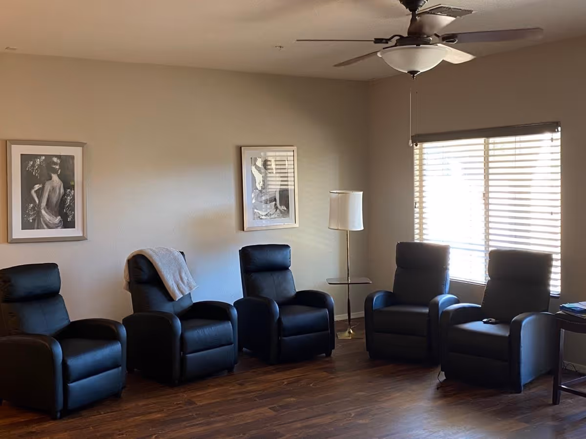 A living room lounge with five black recliner chairs, a lamp, and window blinds.