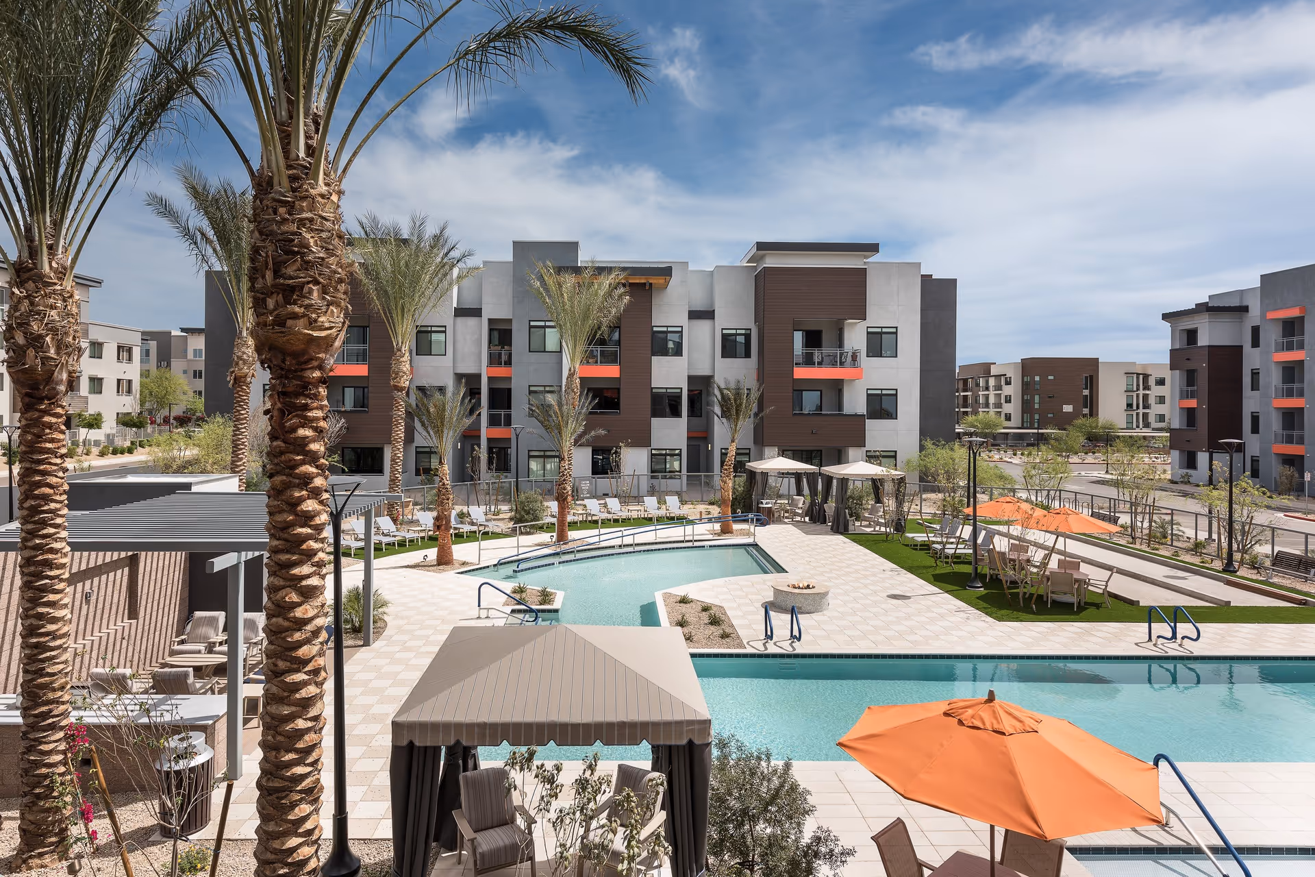 Outdoor view of Clearwater Mayo Blvd facility showing a swimming pool area with lounge chairs, palm trees, shaded cabanas, orange umbrellas, and modern apartment buildings in the background under a partly cloudy sky.