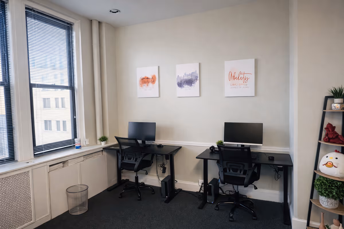 Two desks with computers and chairs in a small office; large windows on the left.