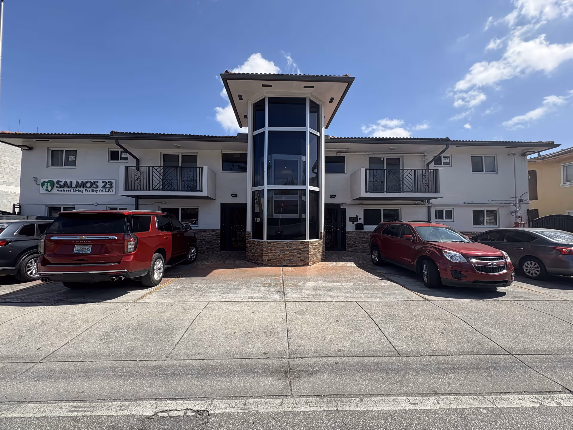Two-story assisted living facility front with a central glass entry tower, balconies, a 'Salmos 23' sign, and several parked cars.