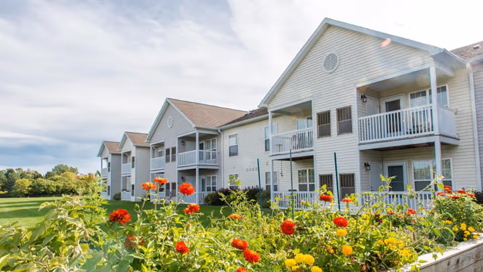 Two-story beige senior living building with balconies, green lawn, and colorful foreground flowers.