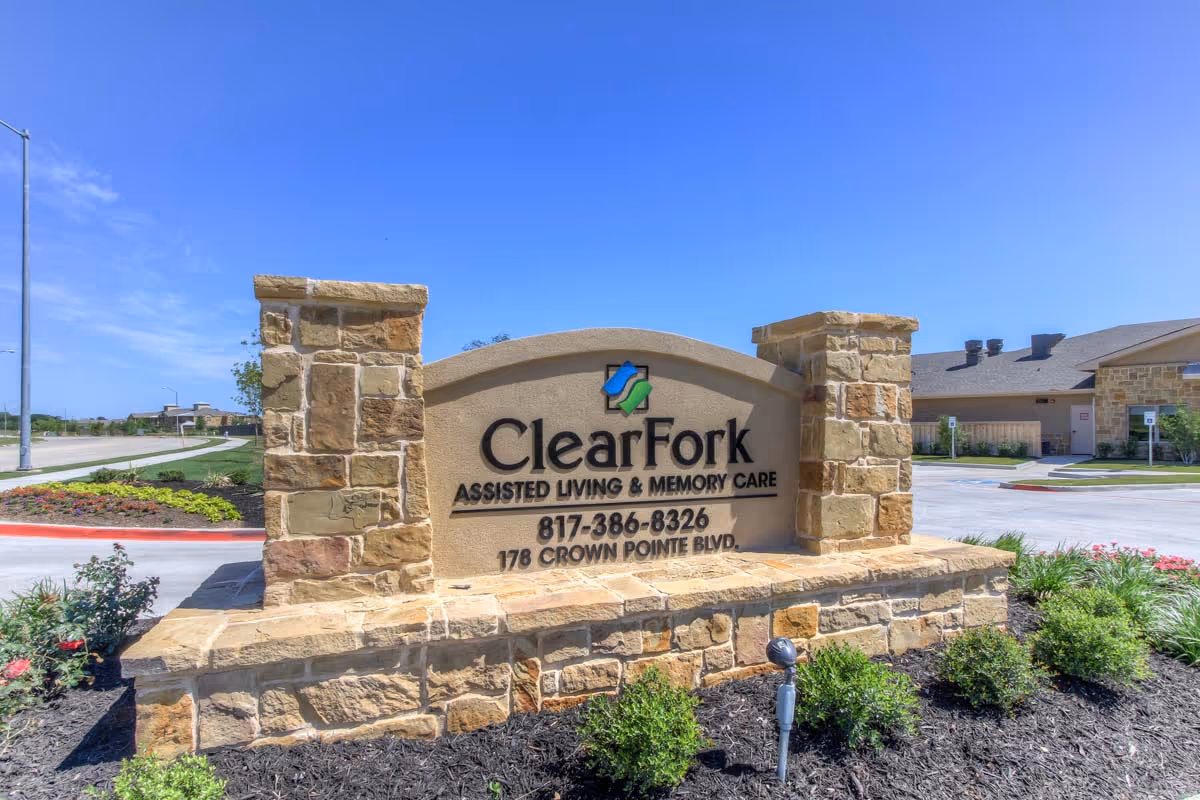 Stone sign at the entrance of Clear Fork Assisted Living and Memory Care facility with the address 178 Crown Pointe Blvd and phone number 817-386-8326, surrounded by landscaping and a clear blue sky.