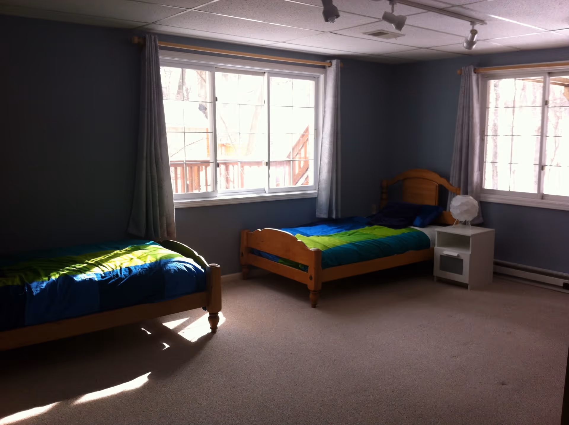 A bedroom with two single wooden beds, each with blue and green bedding. There are two large windows with light gray curtains allowing natural light to fill the room. A small white nightstand with a white spherical lamp is placed between the beds. The walls are painted gray and the floor is carpeted in beige.