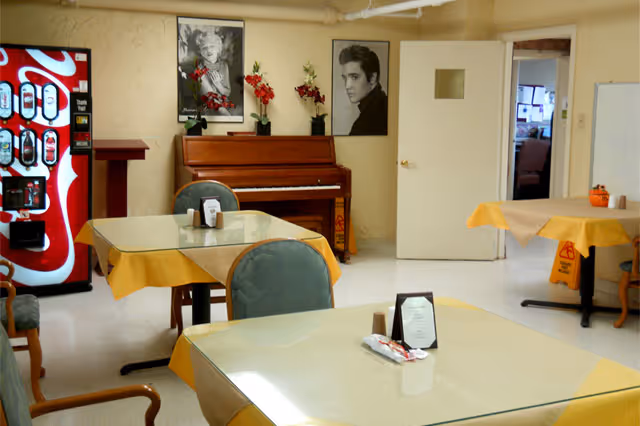 Dining room with tables covered in yellow tablecloths, chairs, a piano against the wall, and a Coca-Cola vending machine.