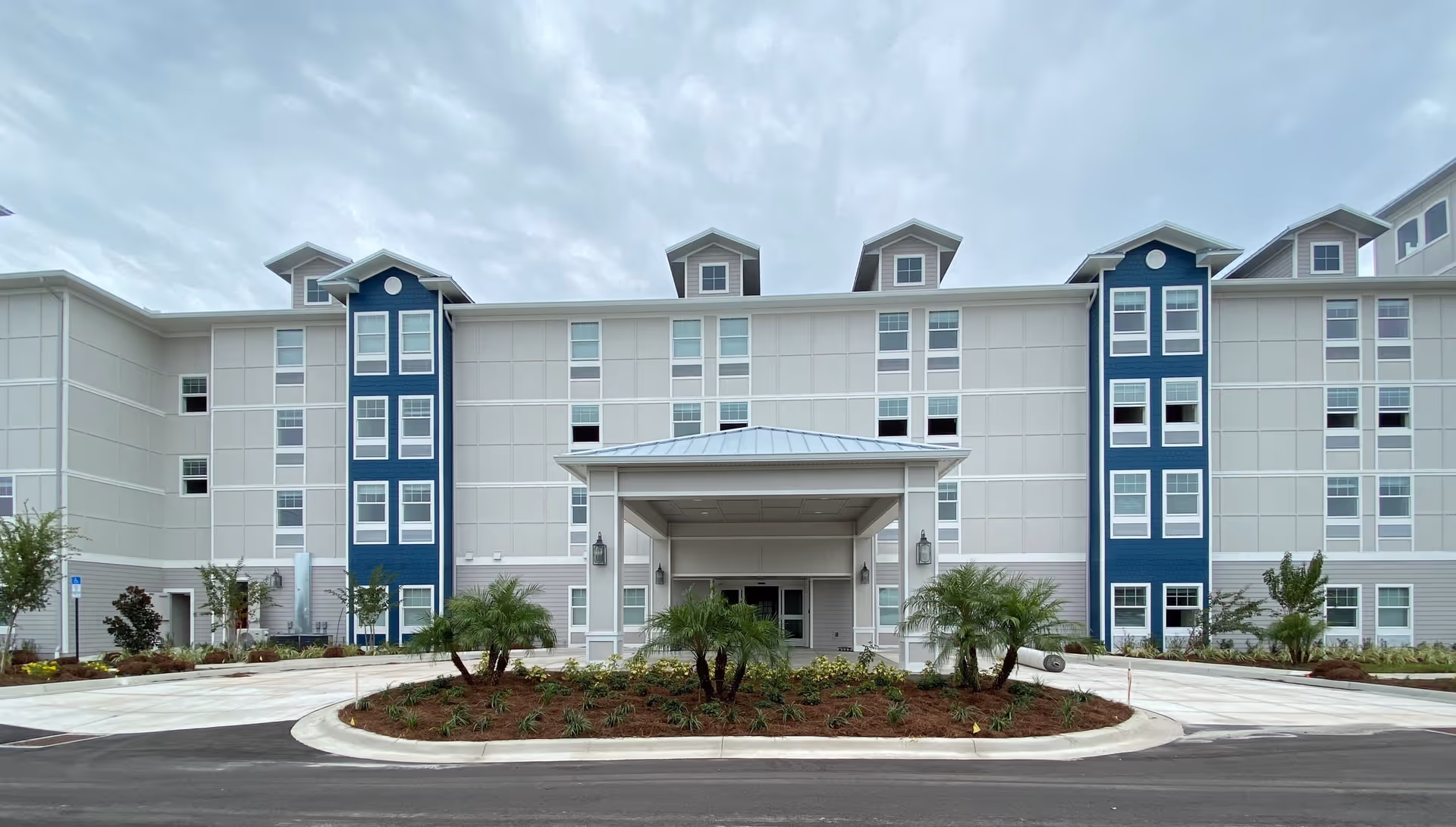 Front entrance of a four-story white and blue senior living building with a covered porte-cochere and landscaped circular driveway.