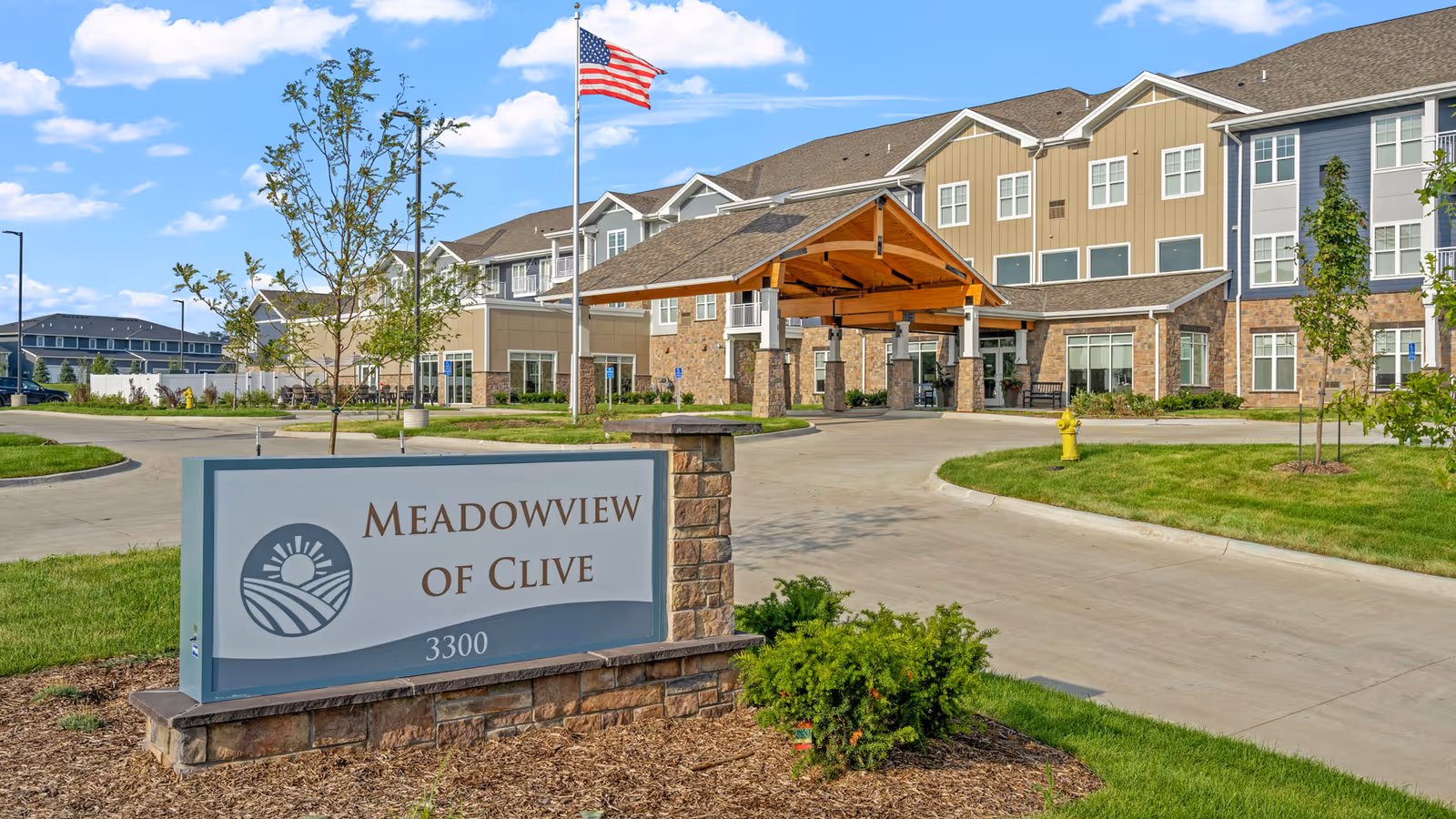 Exterior view of Meadowview of Clive senior living facility showing a large building with multiple windows, a covered entrance with wooden beams, an American flag on a flagpole, landscaped greenery, and a sign with the facility name and address 3300.