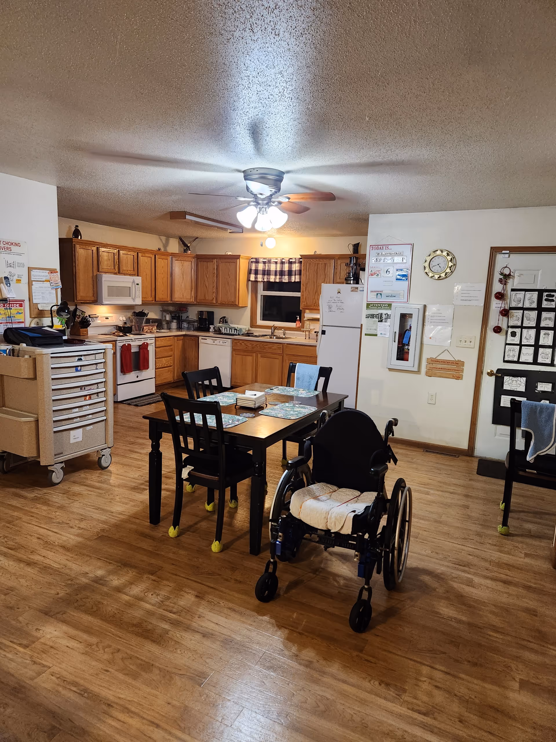 Cozy communal dining area with kitchen, table and chairs, wheelchair, and medical cart visible.