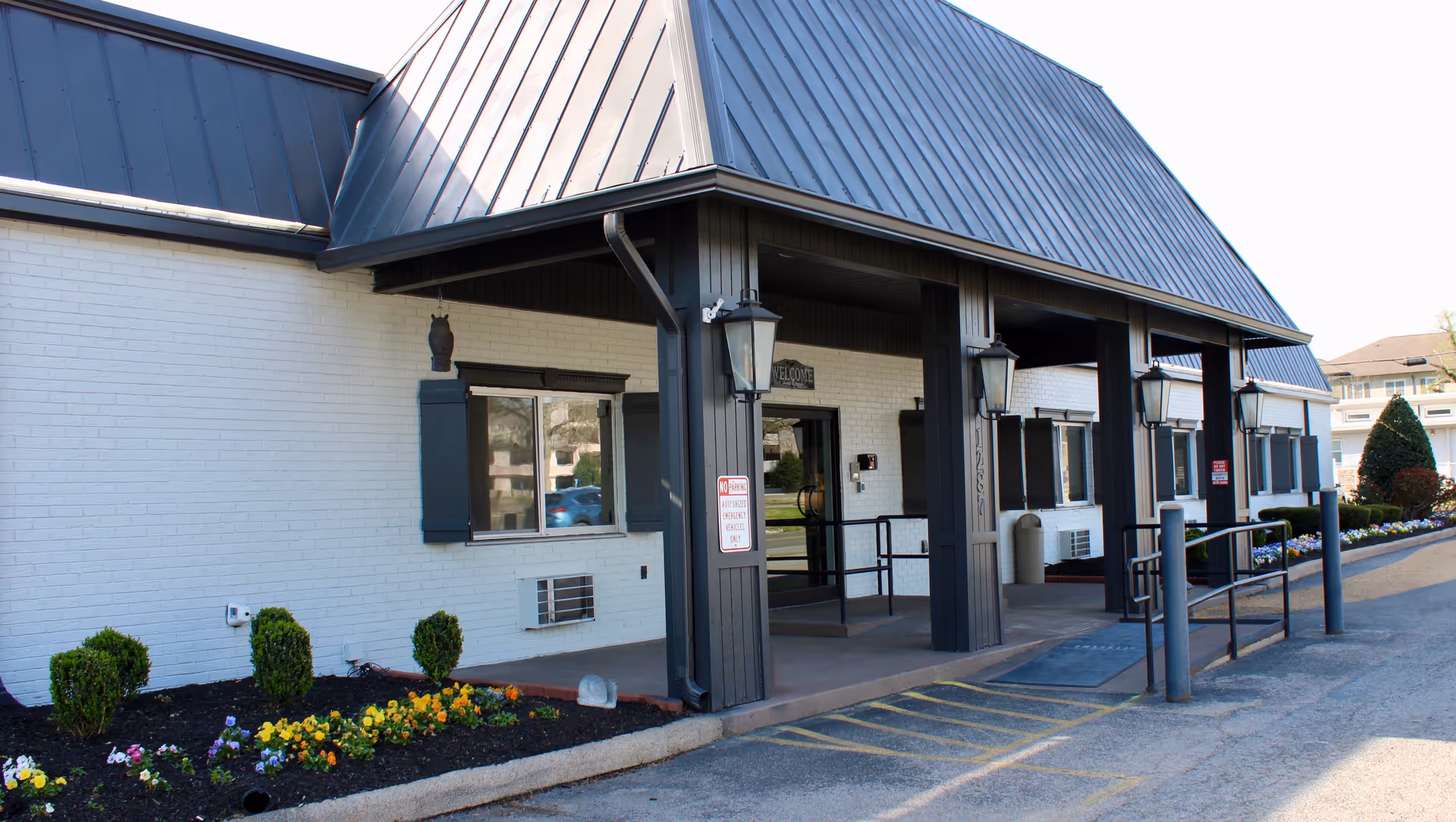 White brick building with dark metal roof, covered entrance, wheelchair ramp, and landscaped flowers.
