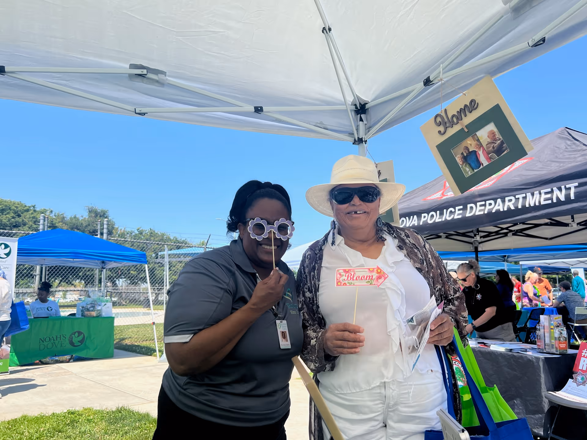 Two smiling people pose under a canopy at an outdoor community fair.
