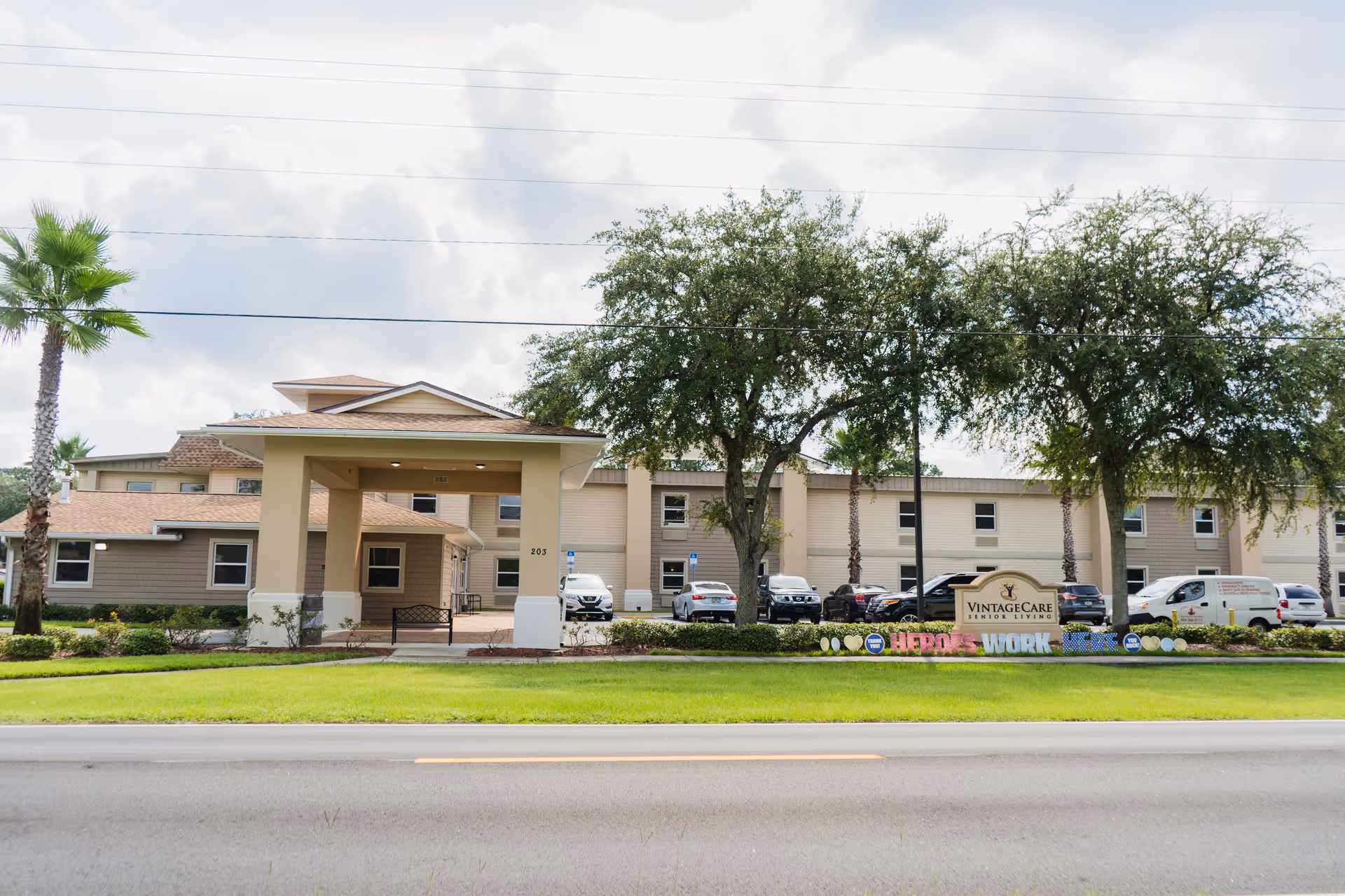 Front exterior view of Vintage Care of Palatka senior living facility with a covered entrance, several parked cars, trees, and a sign displaying the facility name along with a colorful message that reads 'Thank You Heroes Work Here'.