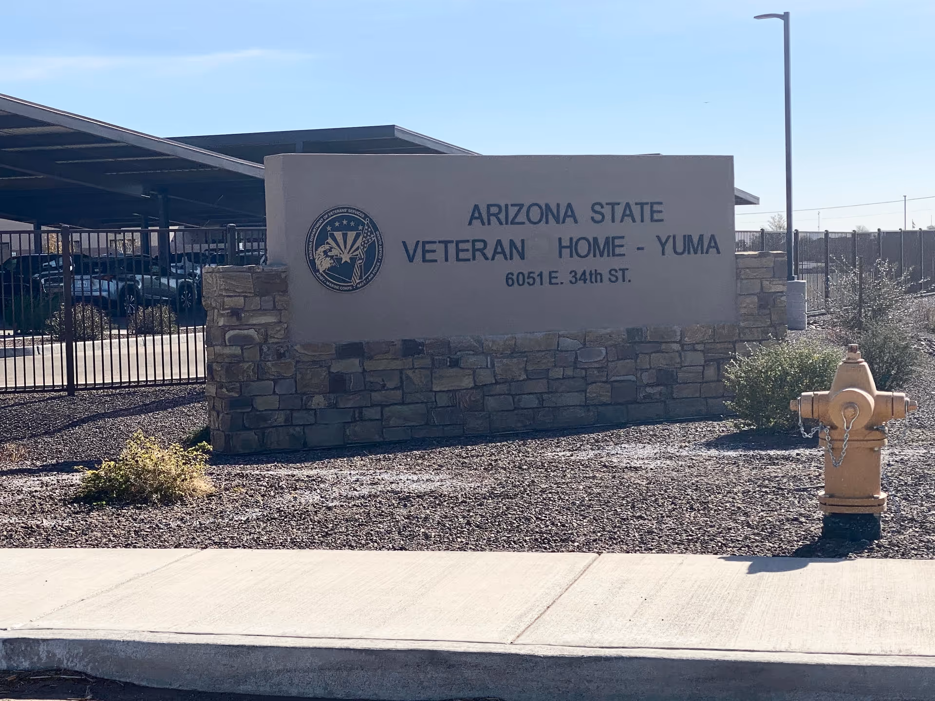 Front exterior sign reads Arizona State Veteran Home Yuma atop a stone wall beside beige hydrant.