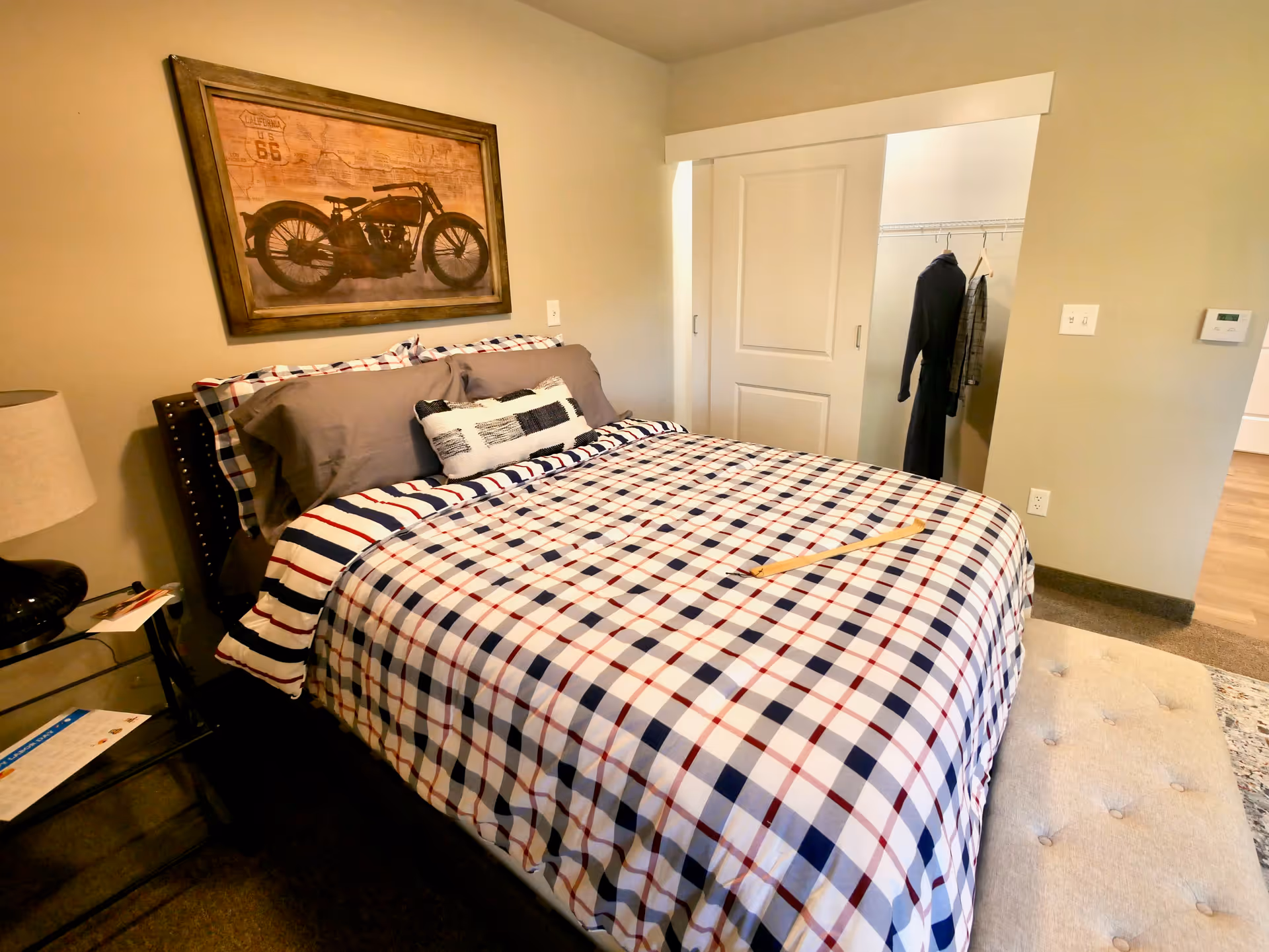 A bedroom with a bed covered in a checkered bedspread in red, white, and blue. There are multiple pillows on the bed, and a wooden cane rests on top. A framed picture of a vintage motorcycle hangs on the wall above the bed. To the right, there is a closet with sliding doors, partially open, revealing clothes hanging inside. A beige cushioned bench is at the foot of the bed, and a small glass side table with a lamp and some papers is on the left side of the bed.