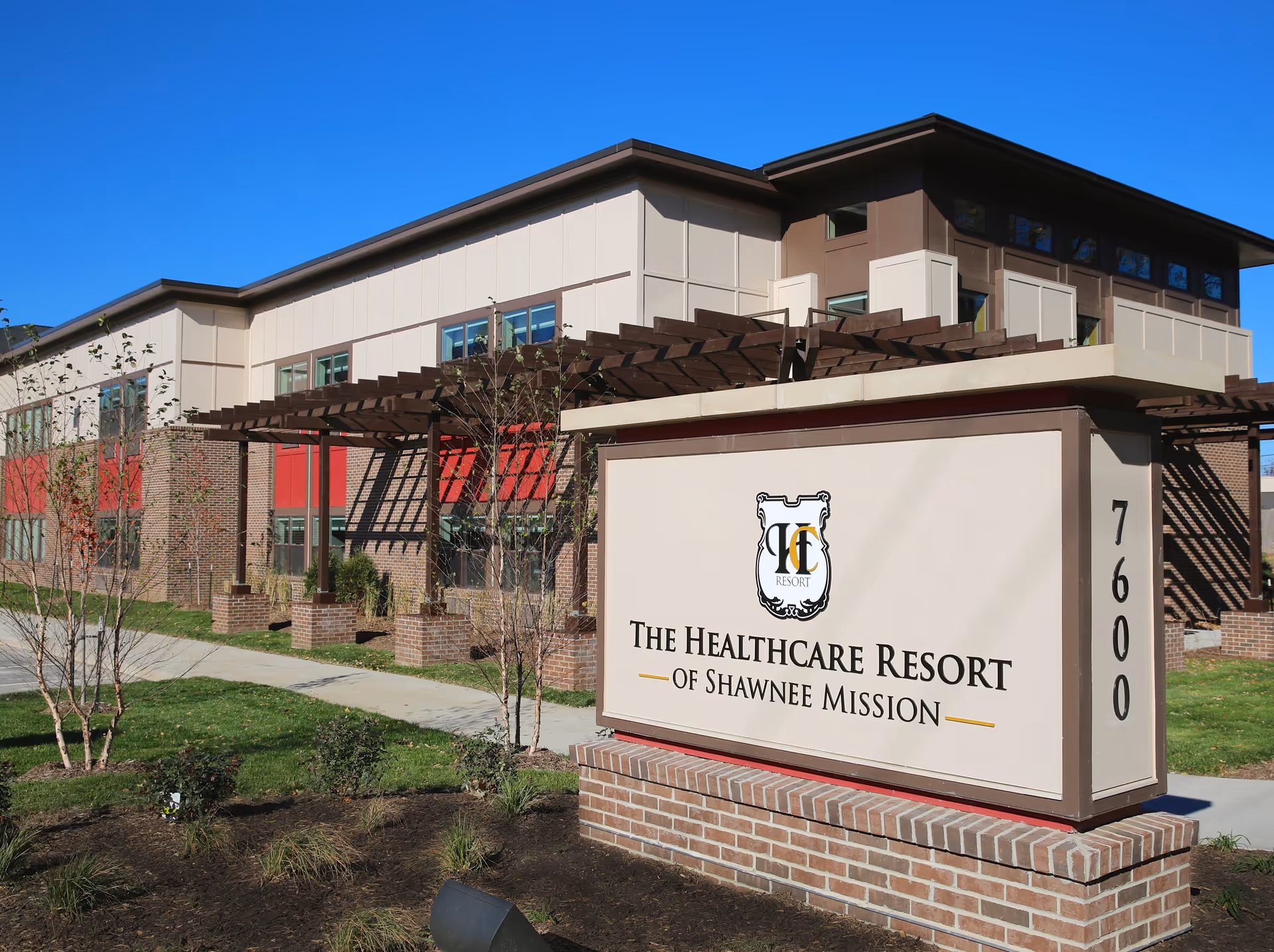 Front exterior of a two-story post-acute rehabilitation building with a large freestanding sign reading "The Healthcare Resort of Shawnee Mission" and the street number 7600.