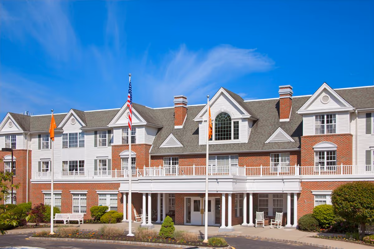 Front exterior of a three-story brick and white senior living facility with flags and a covered entrance.
