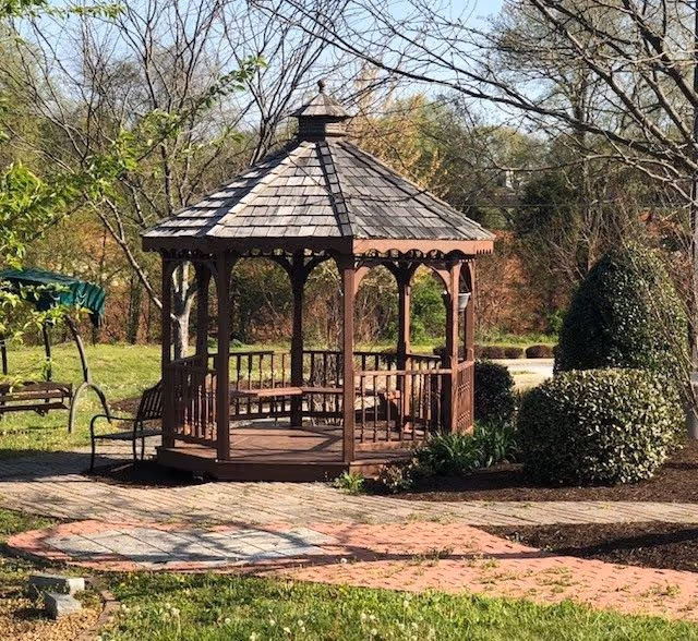A wooden gazebo in a landscaped outdoor garden with brick pathways and trimmed shrubs.