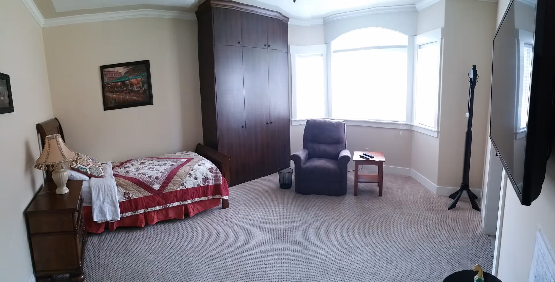 A furnished resident bedroom at Cambridge Senior Care with bed, wardrobe, chair, and side table.