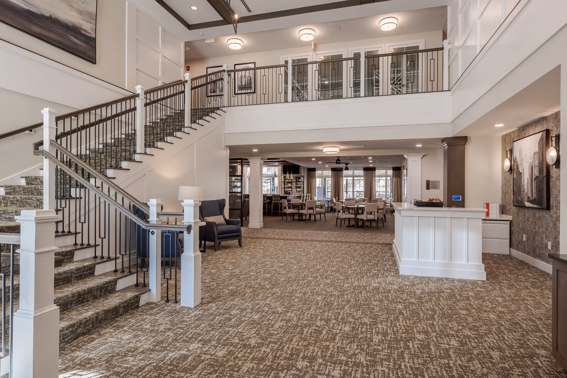 Spacious senior living facility interior lobby with a staircase, reception desk, seating area, and dining area in the background.