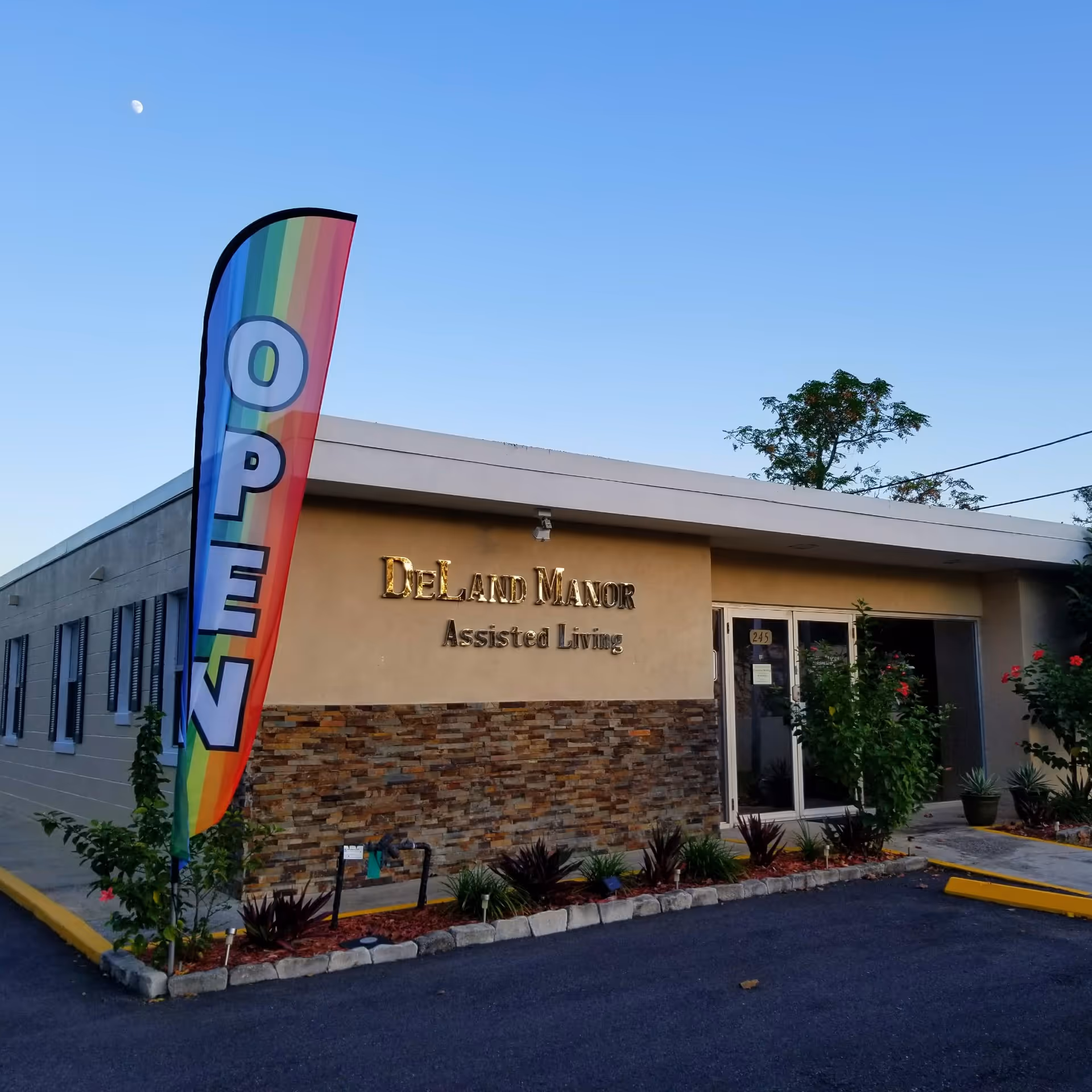 Exterior view of DeLand Manor Assisted Living facility with a rainbow-colored flag displaying the word OPEN. The building has a beige facade with stone accents and some landscaping with plants and flowers near the entrance.