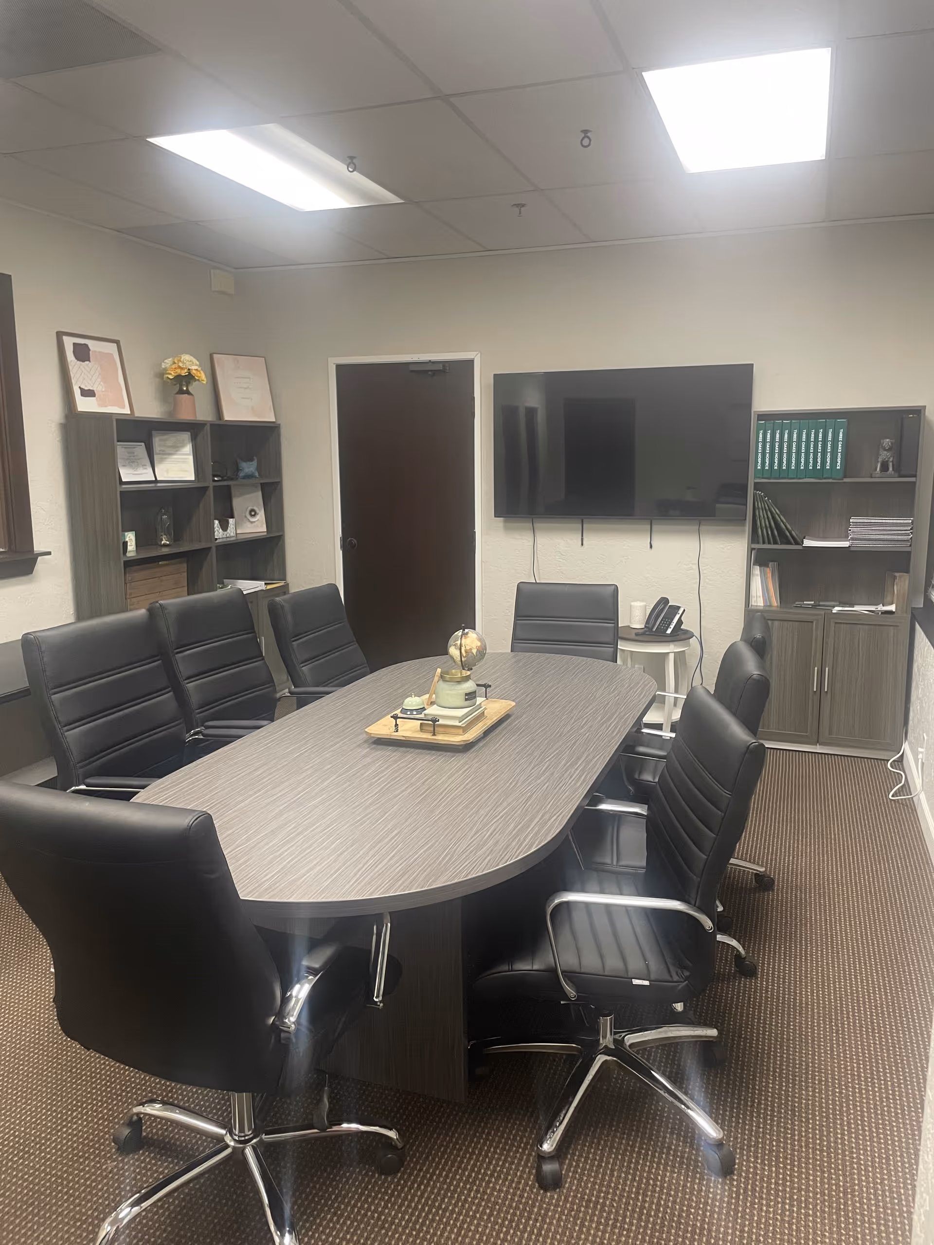 A modern conference room with an oval table, black leather chairs, and a wall-mounted TV.