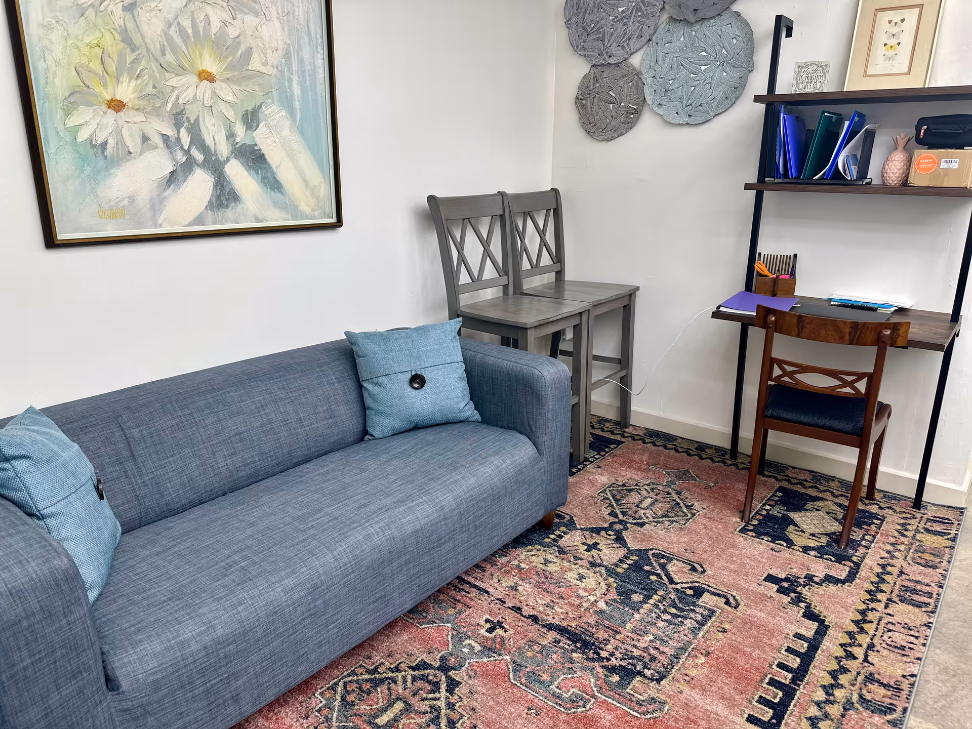 Blue sofa with teal pillows sits beside a desk and stacked chairs in a cozy room.