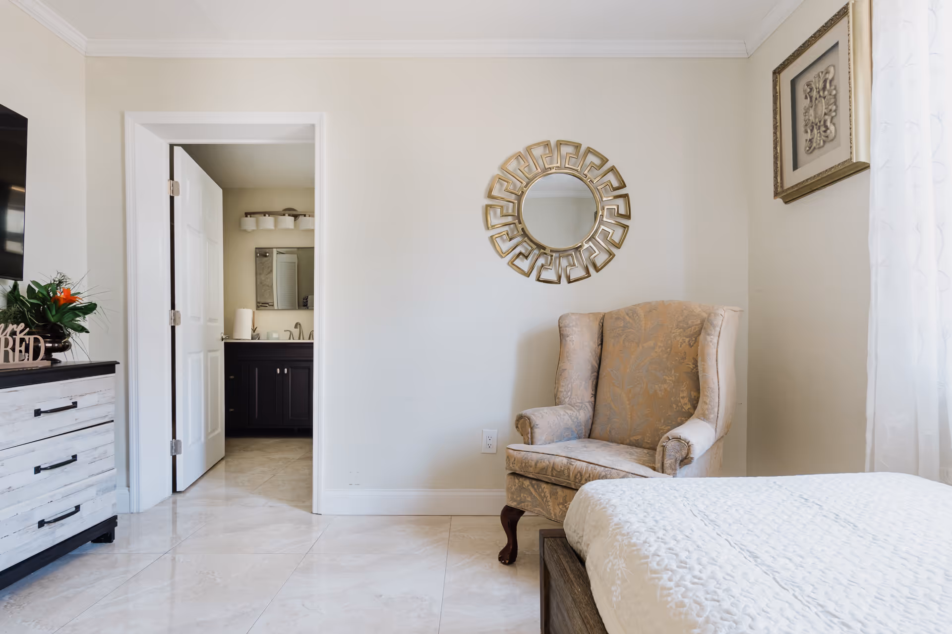 Bright bedroom with a beige armchair, bed in the foreground, a dresser, and an open doorway to a bathroom.