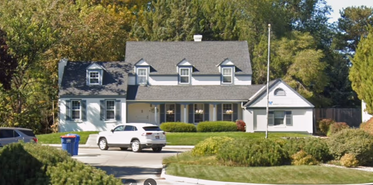 White two-story building with peaked roof, shrubs, and parked cars in front.