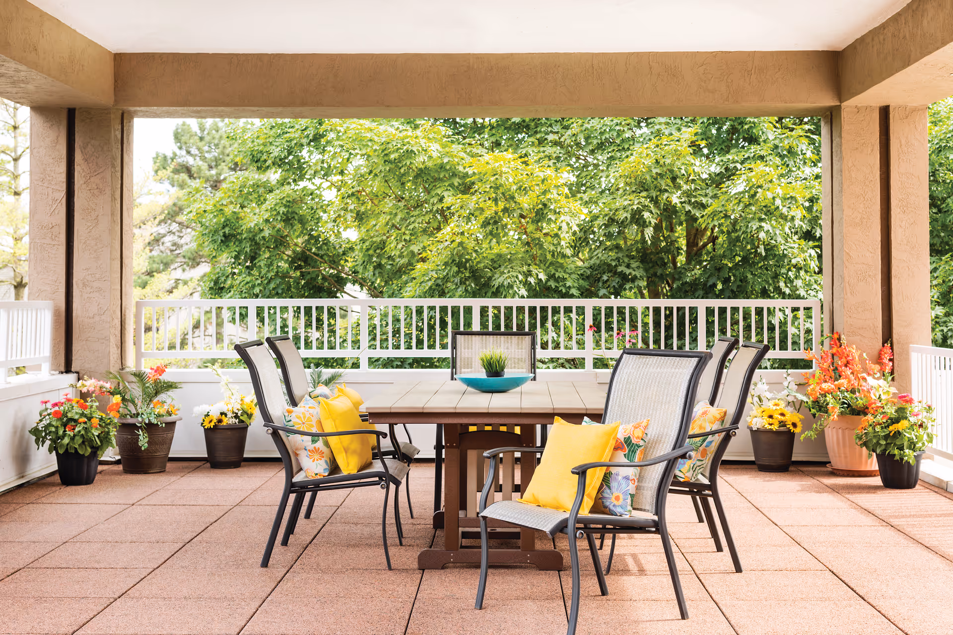 Shaded outdoor patio with a dining table, chairs, and colorful potted flowers.