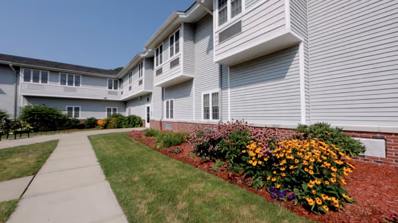 Exterior view of CareOne at Wayne Assisted Living with walkway, lawn, and colorful flower beds.