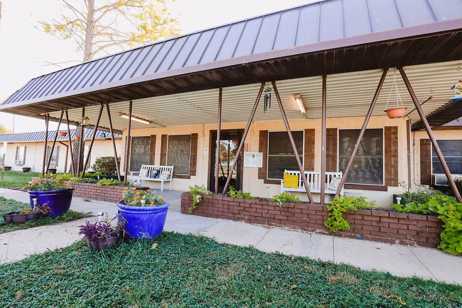Exterior view of Coleman Healthcare Center showing a covered porch area with white benches, potted plants, and brick planters. The building has a metal roof and several windows with brown shutters.