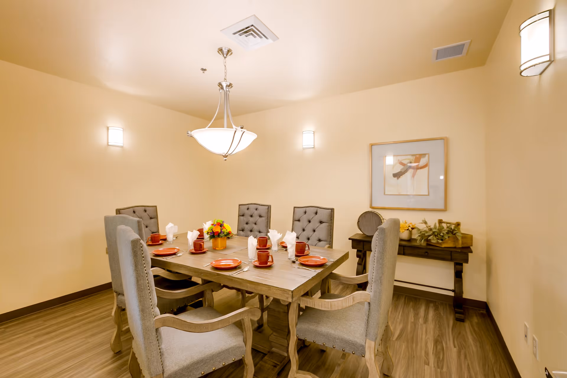 A dining room with a wooden table set for six people. The table has six cushioned chairs with armrests, red plates, matching red cups, and white folded napkins. A small flower arrangement is in the center of the table. The room has beige walls, wooden flooring, a hanging ceiling light, and wall sconces. There is a framed abstract artwork and a small console table with decorative items against the far wall.