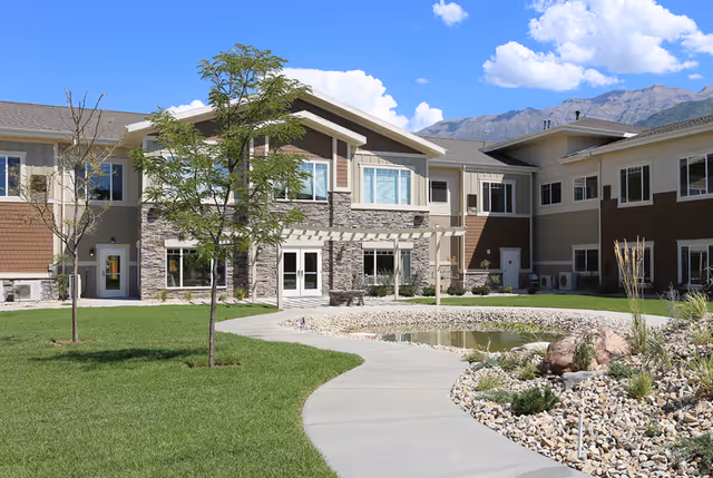 Exterior view of a senior living facility building with a stone and beige facade, surrounded by a well-maintained lawn, young trees, a curved concrete pathway, and a rock garden. Mountains and a blue sky with clouds are visible in the background.