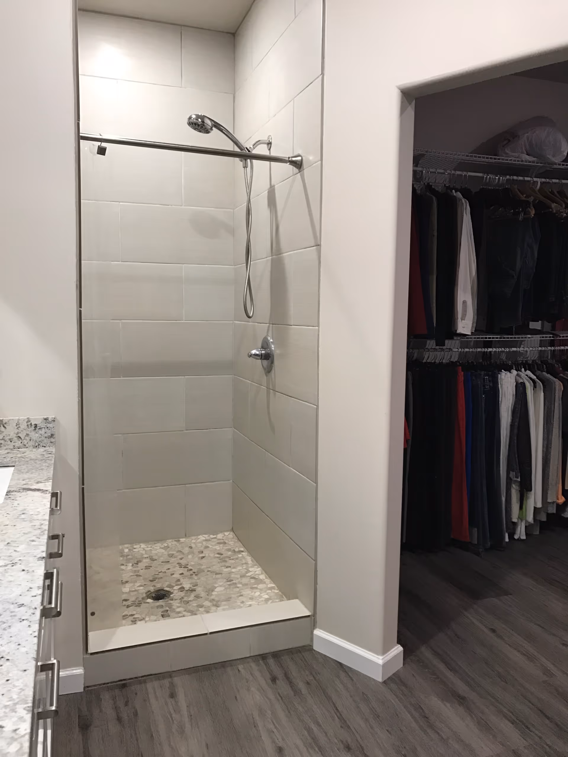 A modern bathroom shower area with light-colored rectangular tiles on the walls and pebble-style tiles on the shower floor. The shower has a chrome showerhead and a control knob. To the right of the shower is an open doorway leading to a walk-in closet with hanging clothes. The bathroom floor is a dark wood-like material, and part of a granite countertop with drawers is visible on the left side.