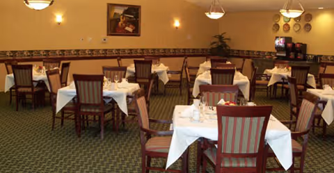 Dimly lit dining room with white tablecloths, wooden chairs, and arranged tables.