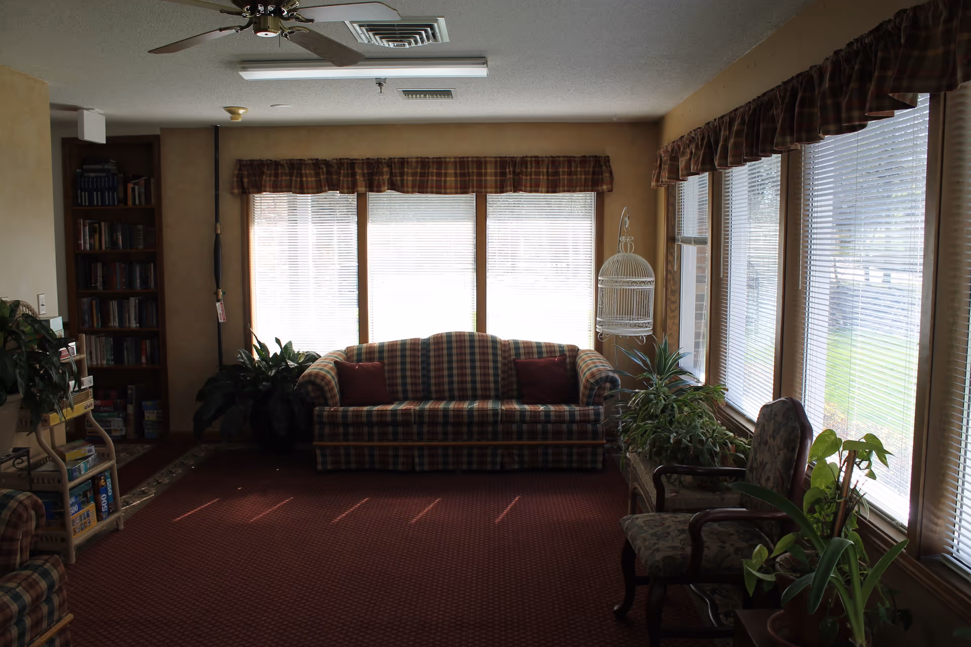 A cozy living room with a plaid sofa and two red pillows in front of large windows with blinds and plaid valances. There are several potted plants, a floral upholstered chair, a white decorative birdcage, and a bookshelf filled with books and board games. The room has a ceiling fan and carpeted floor.