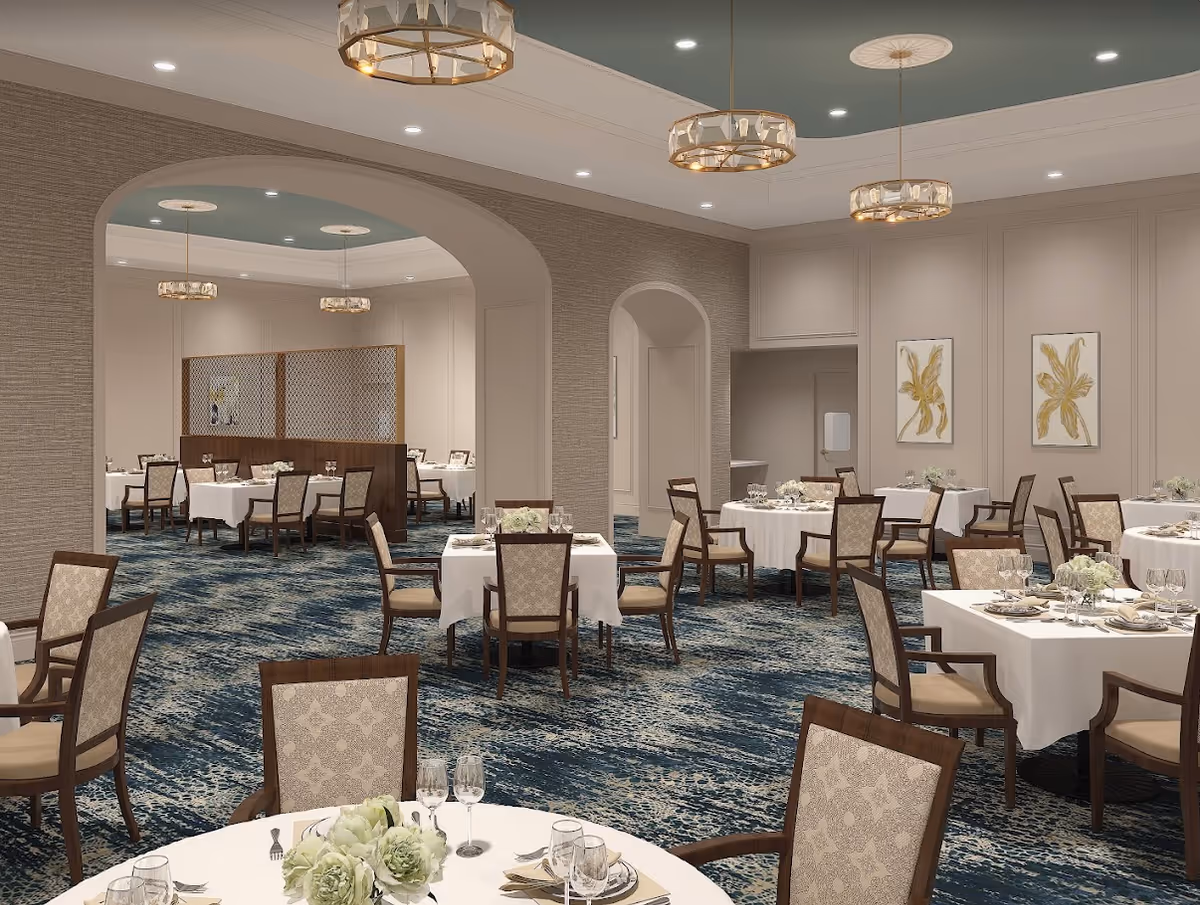 A spacious, elegantly decorated dining room with multiple round and square tables covered with white tablecloths. Each table is set with plates, glasses, silverware, and floral centerpieces. The room features patterned carpet, beige walls with decorative molding, and modern chandeliers hanging from the ceiling. There are archways leading to adjacent dining areas.