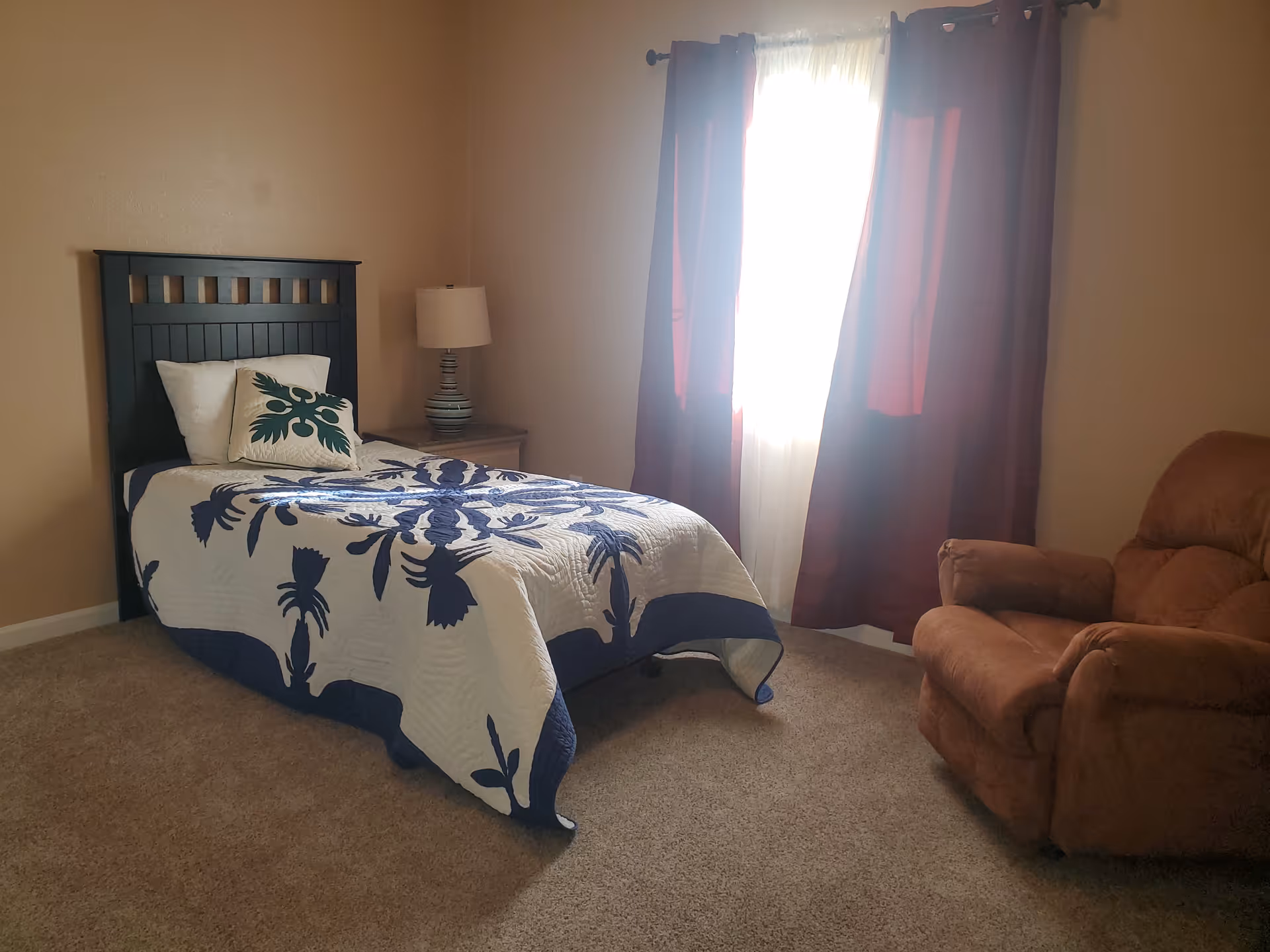 Bedroom with a dark wood bed, nightstand, lamp, window with curtains, and a brown recliner.