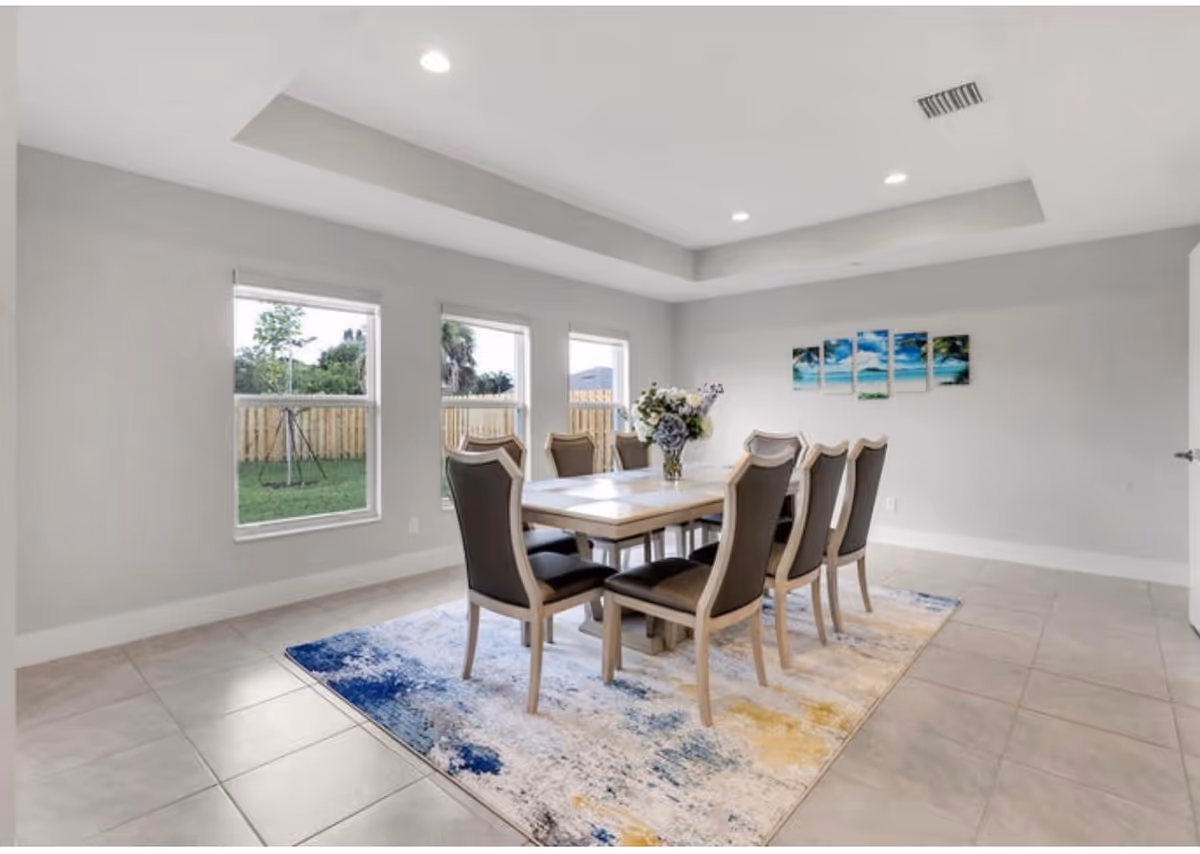 Bright dining room with a long table, eight chairs, large windows, and a decorative rug.