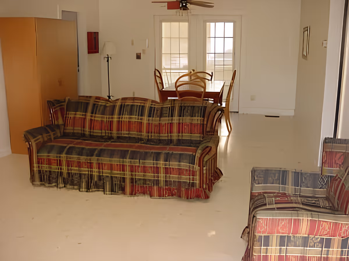 Interior view of a living room with a patterned sofa and loveseat, a wooden dining table with chairs near a set of glass doors, a ceiling fan, and a floor lamp in the corner.