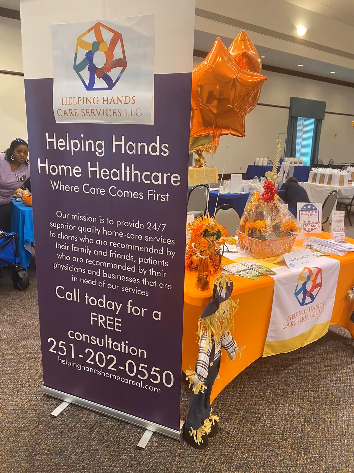 Indoor booth for Helping Hands Care Services LLC with banner, tables, and orange decorations.
