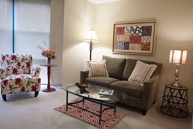 Cozy living room with a sofa, patterned armchair, glass coffee table, lamps, framed artwork, and window blinds.
