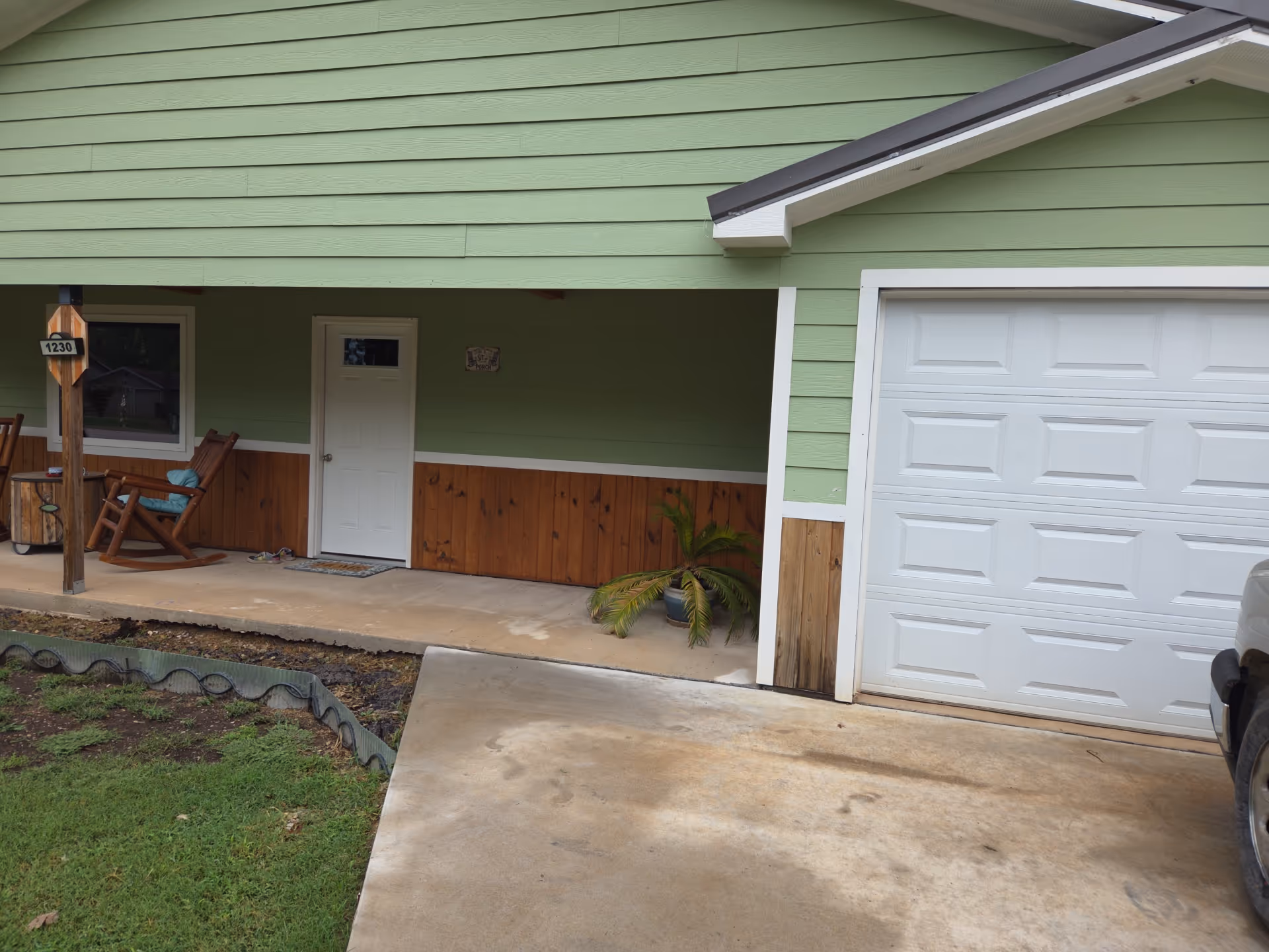 Green house exterior with a covered porch, wooden chairs, and a white garage door.