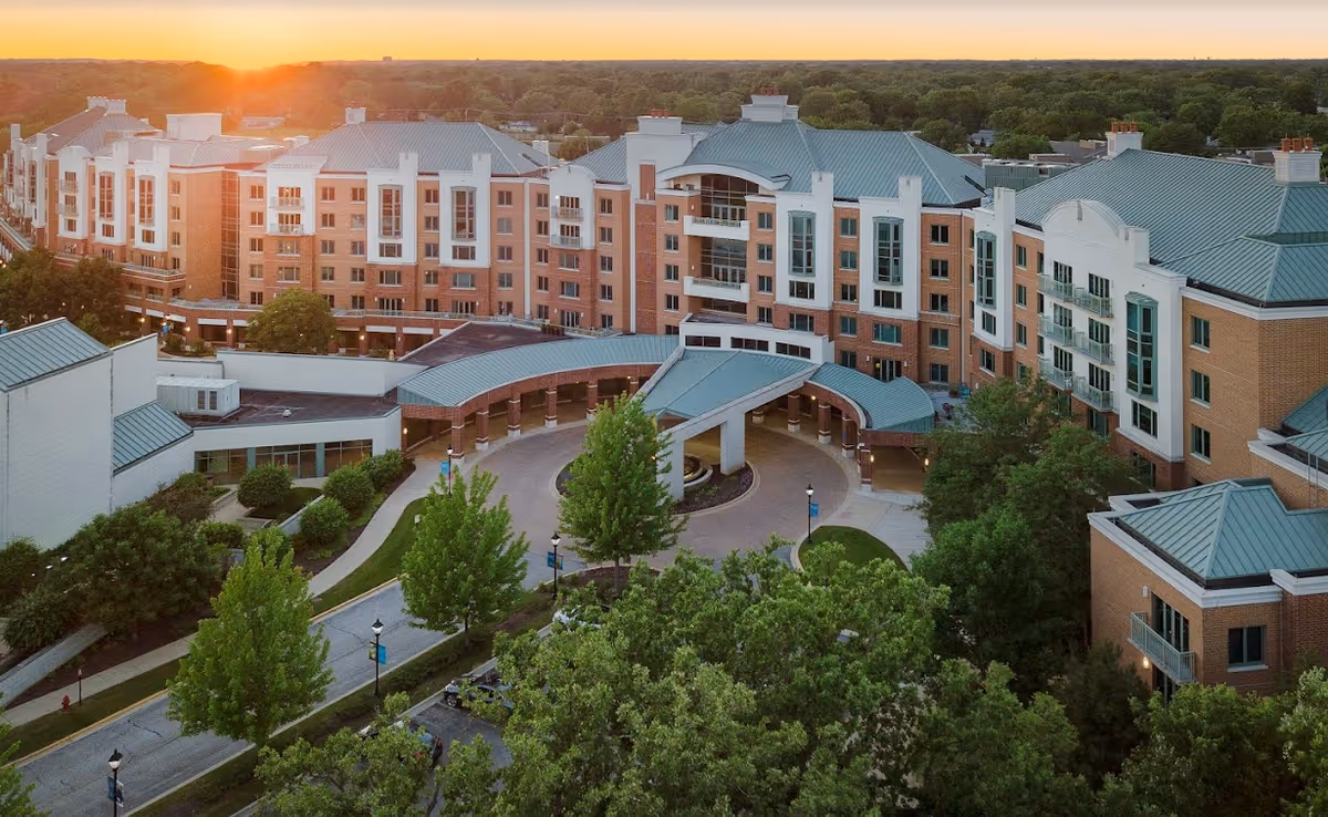 Aerial view of a multi-story brick senior living complex at sunset with a covered circular entrance drive and surrounding trees.