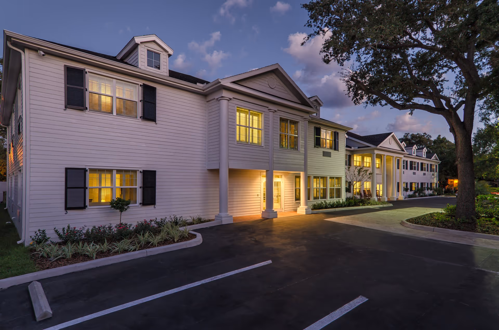 Exterior view of a two-story senior living facility building at dusk with white siding, black shutters, and illuminated windows. The building features large white columns at the entrance and is surrounded by landscaped greenery and a paved parking area.