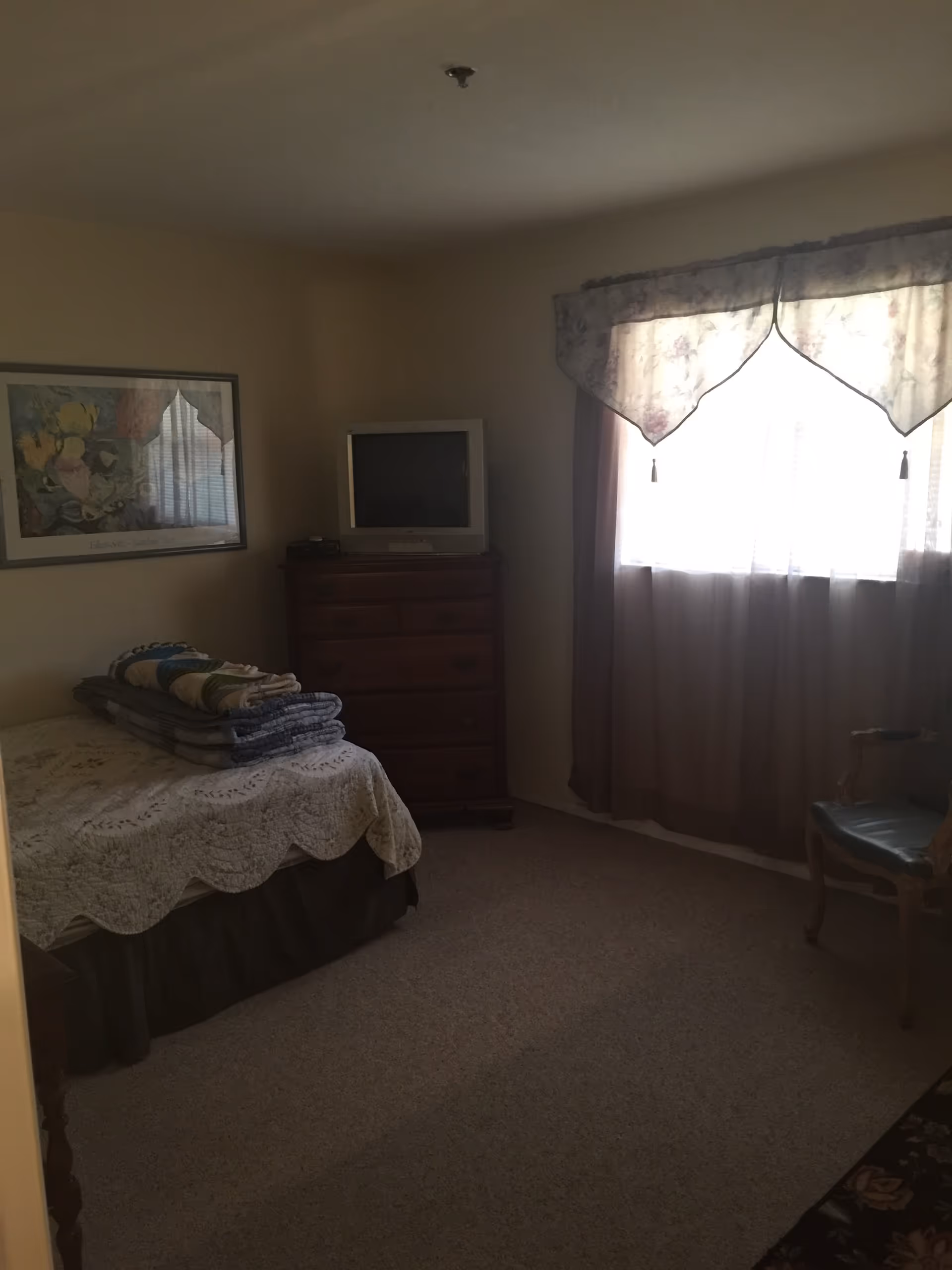 Small bedroom in a senior facility with a bed, dresser, TV, and window curtains.
