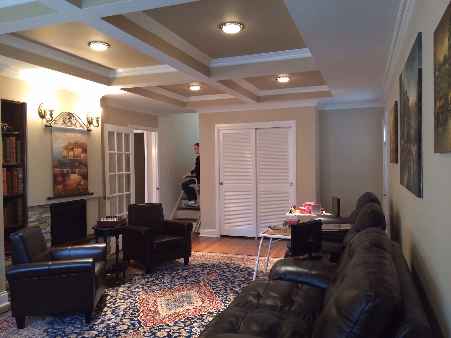A living room with leather sofas and armchairs around a patterned rug, a fireplace, coffered ceiling, and a person on a stairlift in the background.