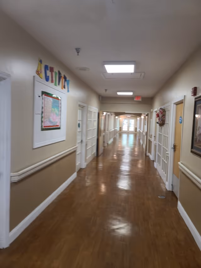 Long polished wood-floored hallway inside a senior living facility with doors, handrails and wall decorations along both sides.